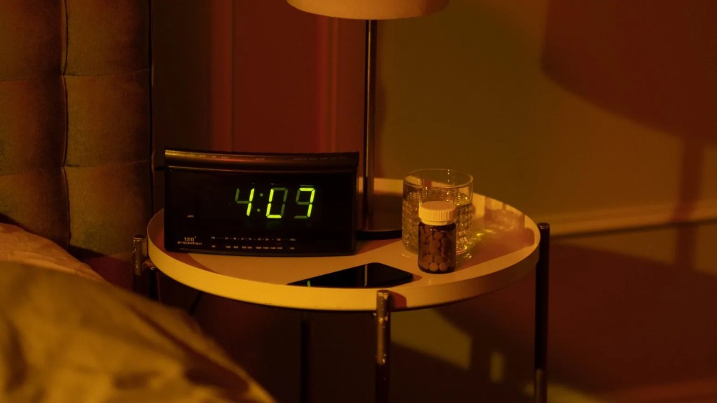 A bedside table at night with a lamp and a digital clock glowing in the early hours.